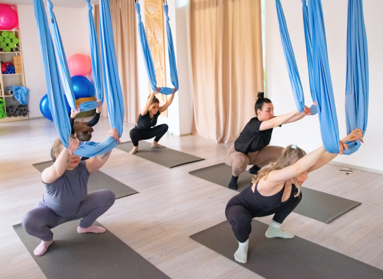 Групповое занятие по направлению «Fly Stretching» в студии «Место Силы» в Батуми: участницы выполняют глубокие приседания, используя подвесные гамаки для поддержки и баланса